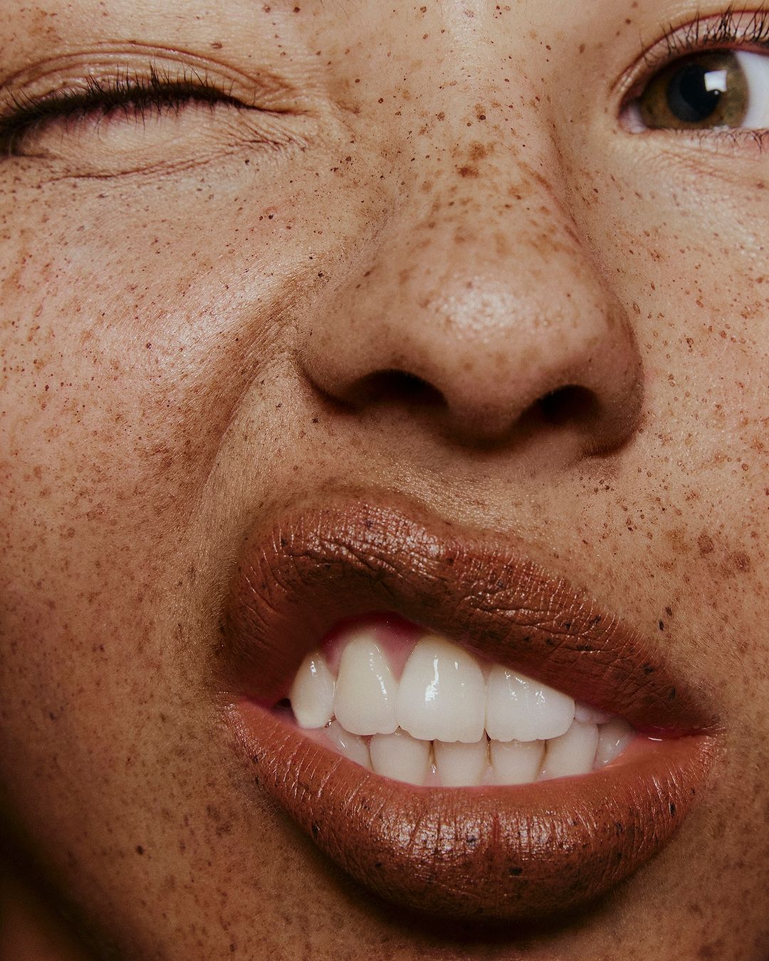 Close-up portrait of a woman with freckles, one eye closed, and a playful expression, wearing brown lipstick with glossy sheen. 