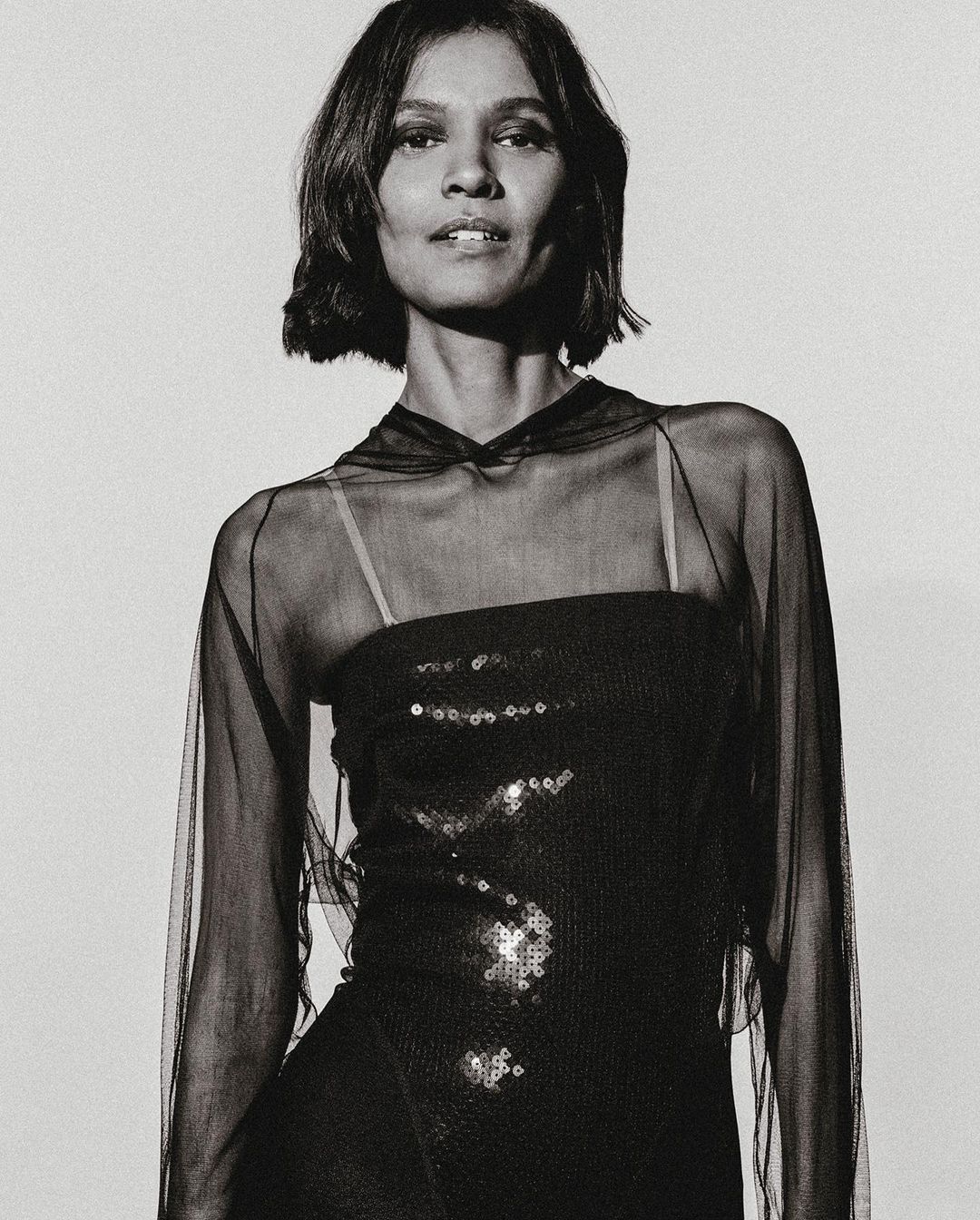 Monochrome portrait of a woman with short dark hair wearing a sequined black dress and a sheer black hooded cape in a photo studio.