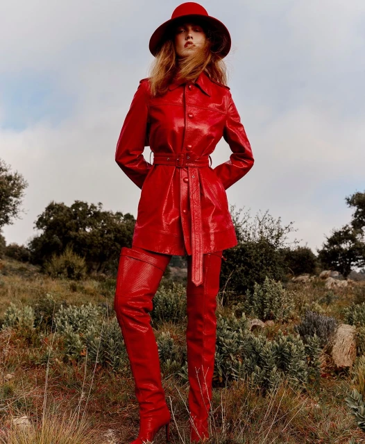 Adult female in red patent leather hat, coat, and thigh-high boots stands in a field of high grass with cloudy skies in the background.