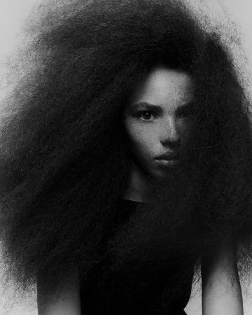 Portrait of a woman with large voluminous textured hair and freckles against a neutral background by Charlotte Navio