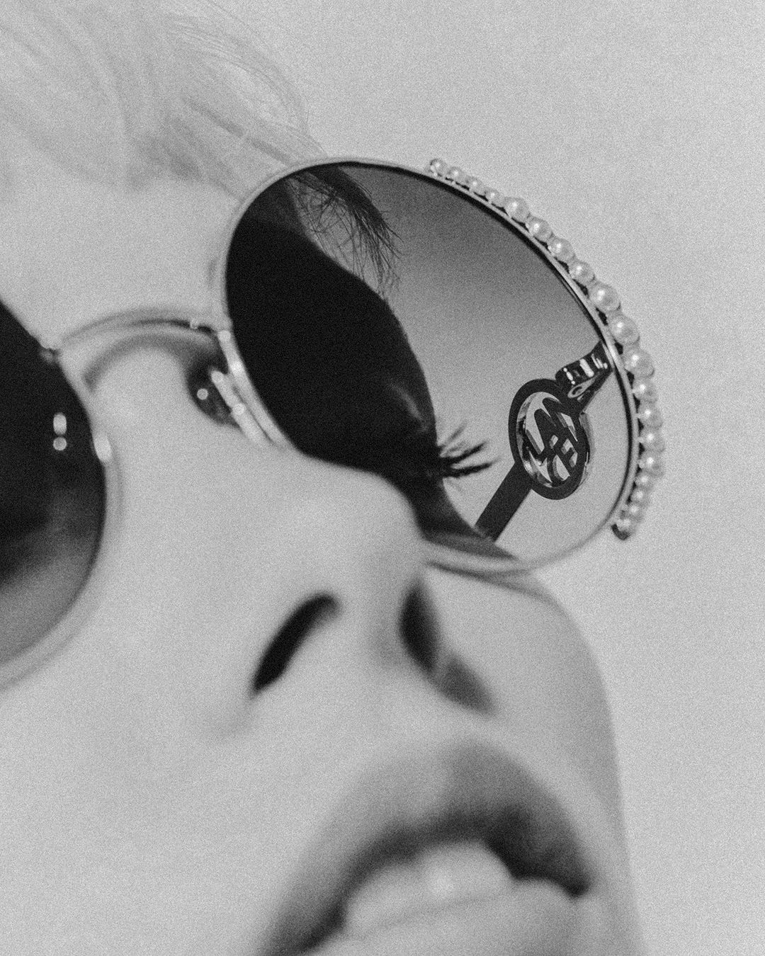 Close-up black and white shot of a person wearing pearl-embellished sunglasses. The sunglasses have a reflected logo in the lens.