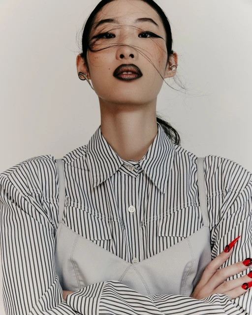Close-up portrait of an Asian woman with bold makeup, a striped shirt, and red nails in a light indoor studio environment.