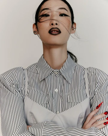 Close-up portrait of an Asian woman with bold makeup, a striped shirt, and red nails in a light indoor studio environment.