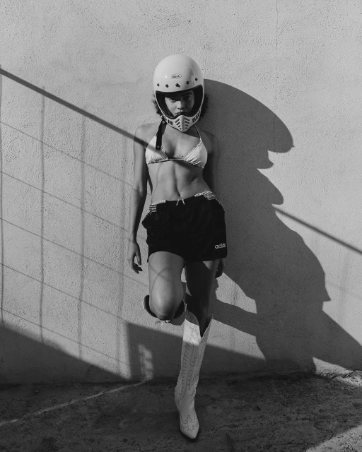 A woman in a bikini, shorts, and helmet leans against a wall, lit by shadows. She's wearing cowboy boots in black and white.