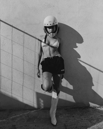 A woman in a bikini, shorts, and helmet leans against a wall, lit by shadows. She's wearing cowboy boots in black and white.