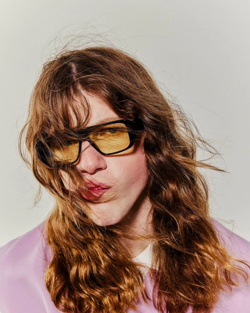 Headshot of a young adult with long brown hair, wearing yellow-tinted sunglasses and a lavender jacket against a light background.