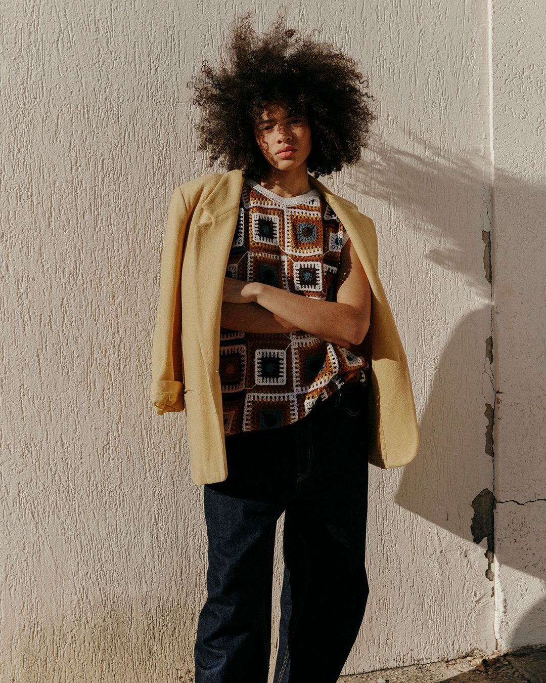 Portrait of an adult with large curly hair, wearing a patterned top with a blazer over their shoulder standing against a stucco wall.