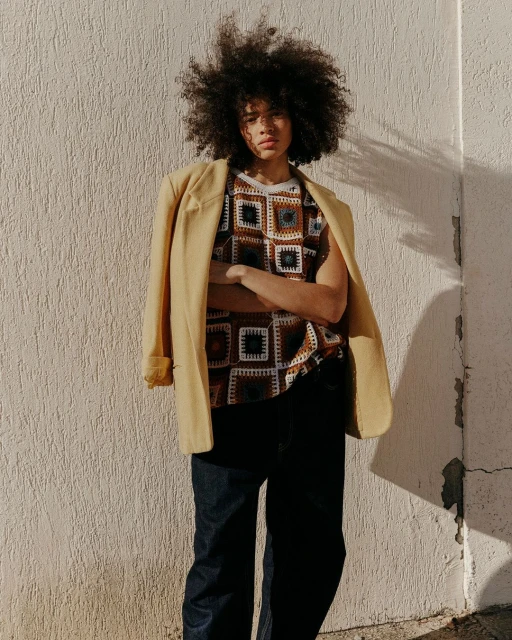 Portrait of an adult with large curly hair, wearing a patterned top with a blazer over their shoulder standing against a stucco wall.