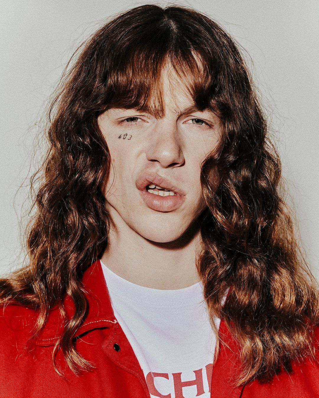 Close-up portrait of a young adult with long brown hair and a red jacket, showcasing a unique facial expression against a neutral background.