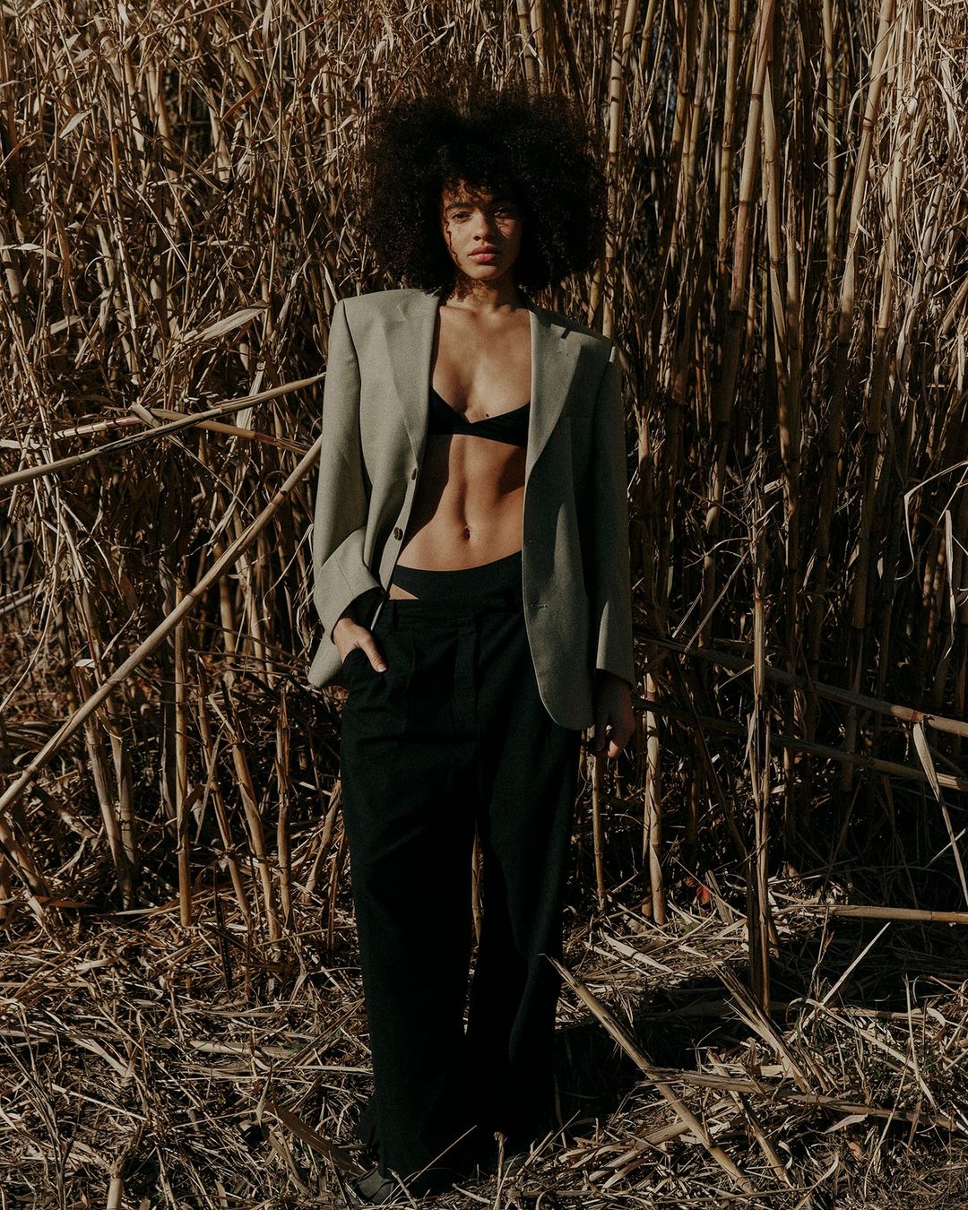 A young mixed-race woman poses outdoors wearing a blazer, bra top, and dress pants, against a background of tall dry grass.