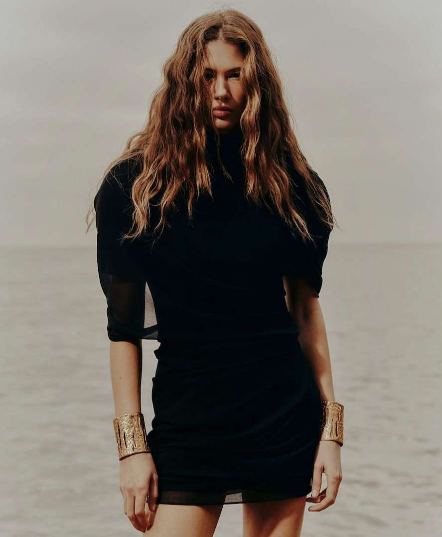 A woman with long wavy hair stands on a beach wearing a black dress and golden bracelets, with a neutral expression, against an ocean backdrop.
