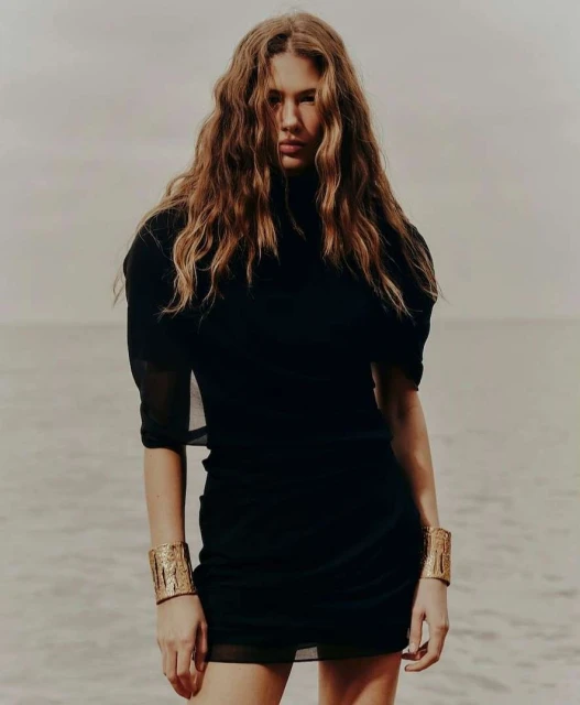 A woman with long wavy hair stands on a beach wearing a black dress and golden bracelets, with a neutral expression, against an ocean backdrop.