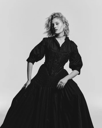 Portrait of a blonde woman wearing a black period dress and necklace against a seamless grey backdrop