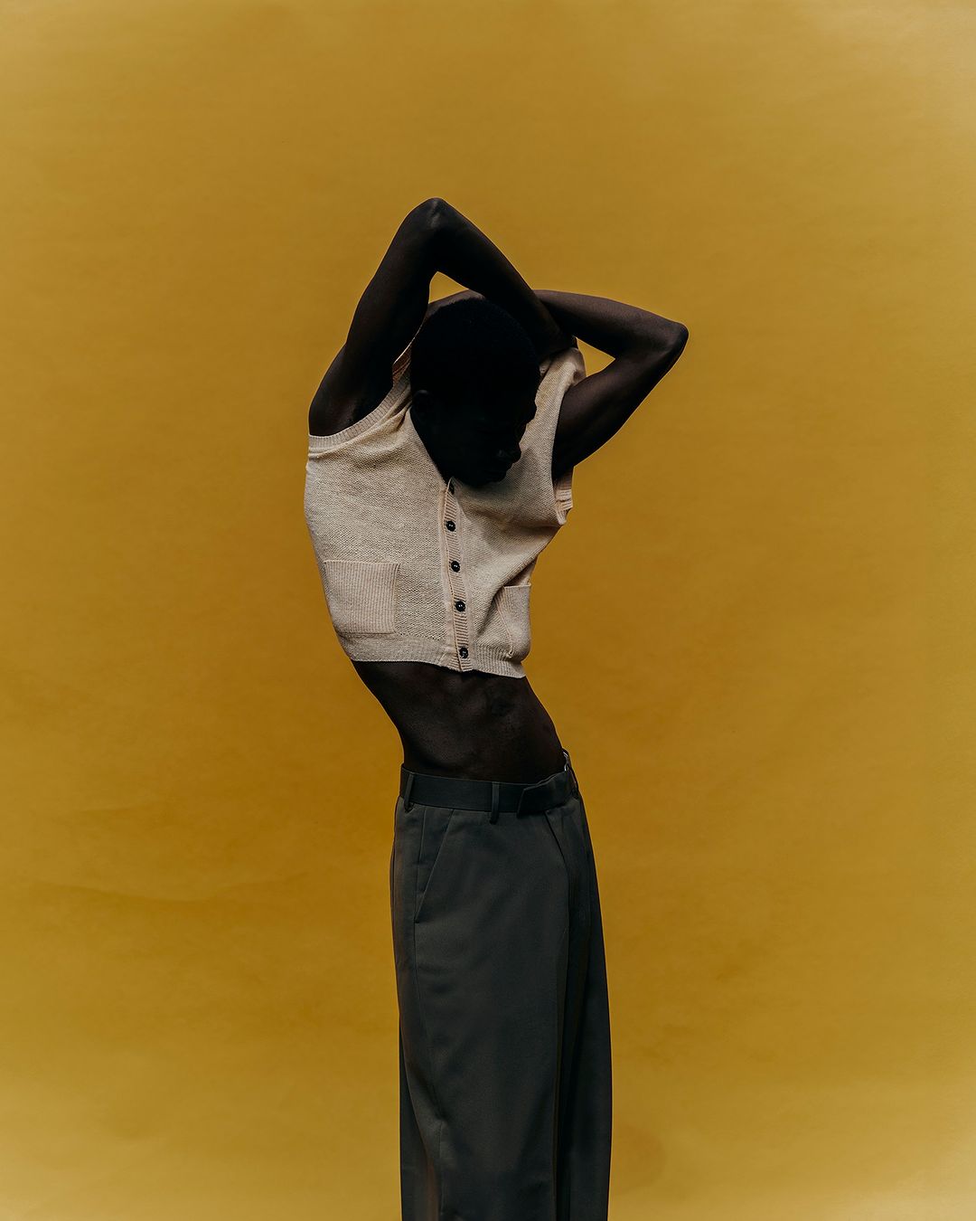 A dark-skinned adult poses against a solid yellow backdrop wearing a knit top and grey pants, with their hands behind their head.