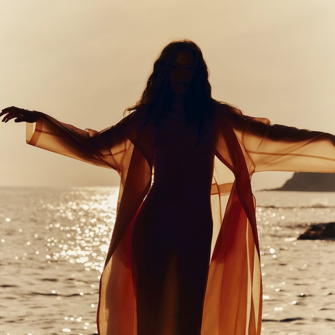 Silhouette of a woman in a flowing orange robe standing on a rocky shore, facing the sea with arms outstretched at sunset.