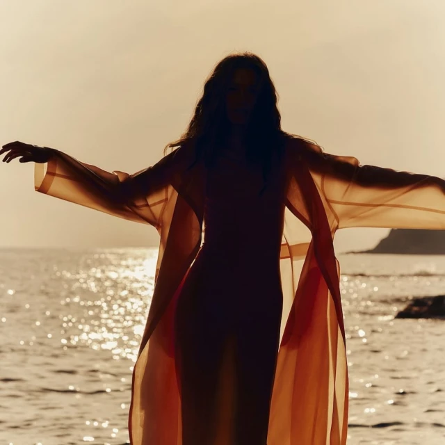 Silhouette of a woman in a flowing orange robe standing on a rocky shore, facing the sea with arms outstretched at sunset.