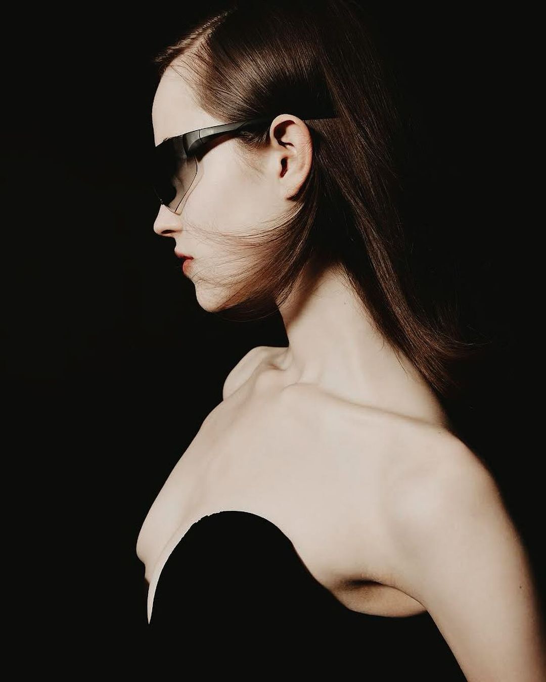 Profile portrait of a woman with dark hair wearing futuristic sunglasses against a black background in a high-fashion beauty shot.