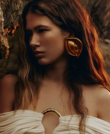 Close-up portrait of a woman with long wavy brown hair wearing large gold earrings and an off-shoulder white top outdoors