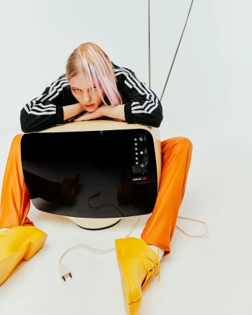 Woman with blond hair, wearing an Adidas track jacket and orange pants, leans against a vintage TV in a minimalist white studio.