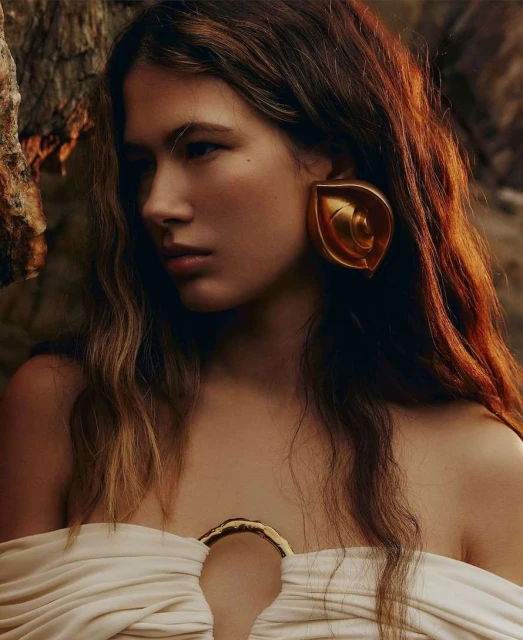 Close-up of a young woman with wavy brown hair, wearing a white dress and large gold earrings, posing outdoors near rocky terrain.
