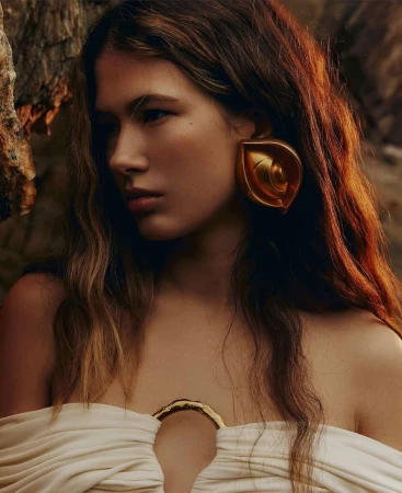Close-up of a young woman with wavy brown hair, wearing a white dress and large gold earrings, posing outdoors near rocky terrain.