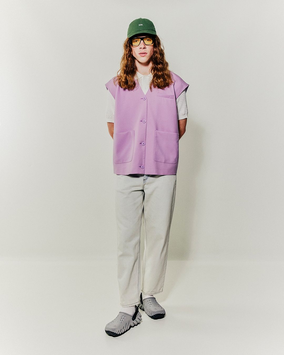 Model with long wavy hair wearing a lavender vest, green cap, and light pants against a plain white background