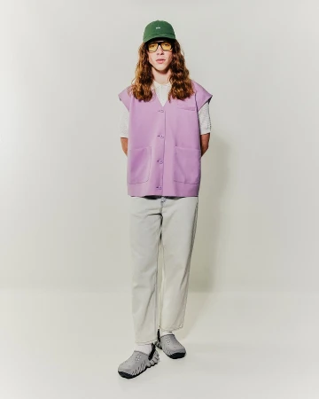Model with long wavy hair wearing a lavender vest, green cap, and light pants against a plain white background