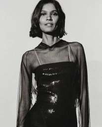 Monochrome portrait of a woman with dark short hair wearing a sheer black top and sequined dress against a pale background.