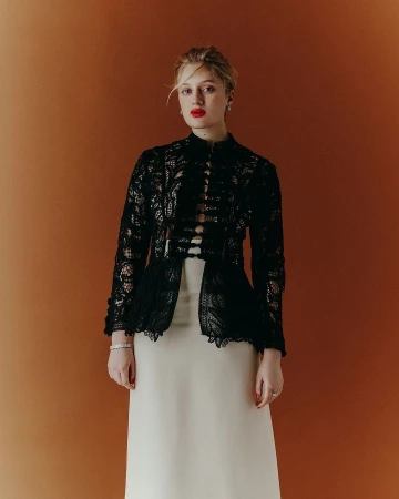 Model wearing a black lace jacket over a light cream skirt against an orange-brown backdrop, photographed by Charlotte Navio.