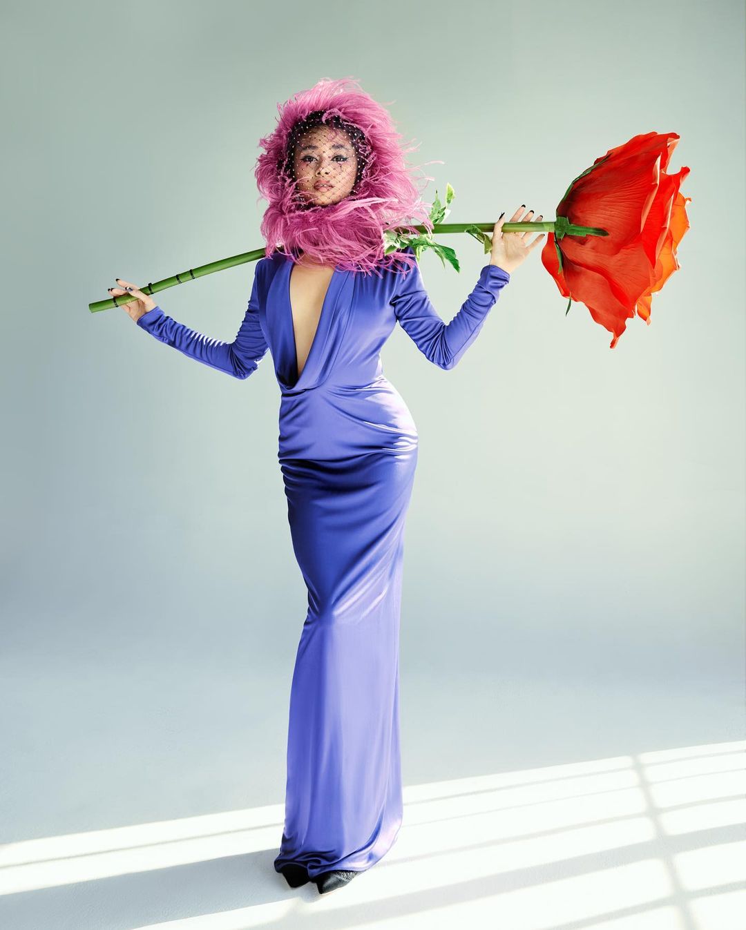 A woman in a periwinkle gown with a pink feathered headpiece holds a giant red rose against a light gray background in a studio shot.