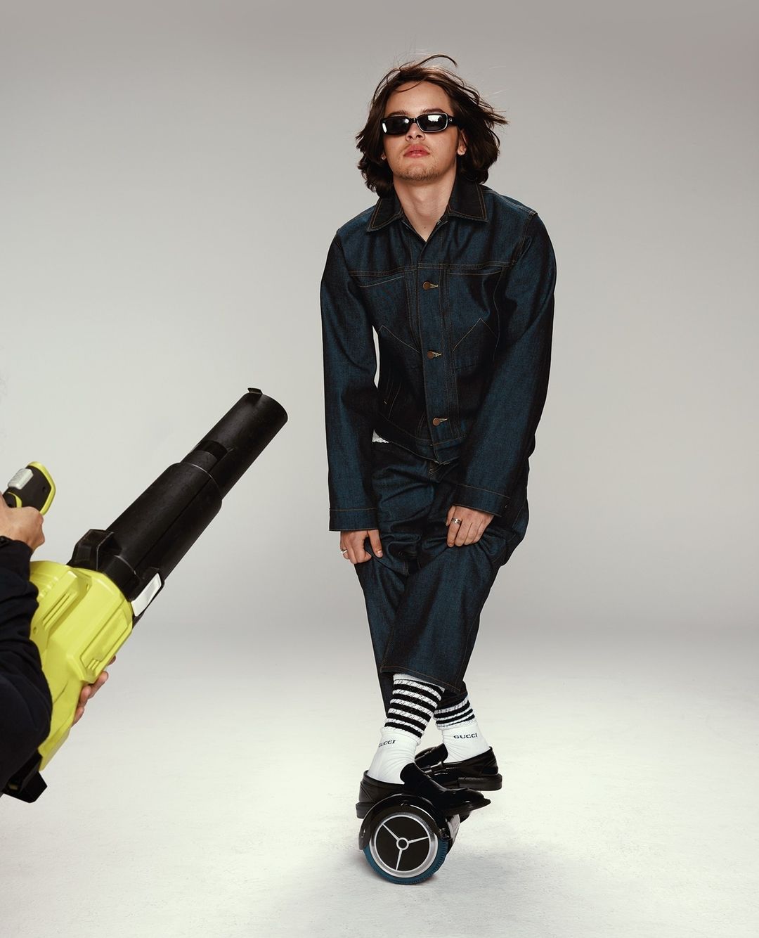 Man in denim jumpsuit and sunglasses rides a hoverboard as someone uses a leaf blower on him in a studio setting against a grey backdrop.