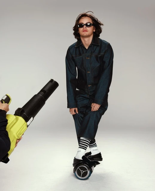Man in denim jumpsuit and sunglasses rides a hoverboard as someone uses a leaf blower on him in a studio setting against a grey backdrop.