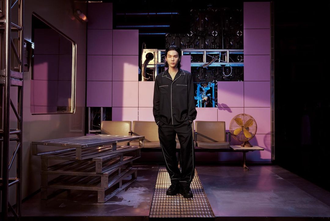 An adult male stands indoors wearing dark pajamas with white piping. Stage setting with metal grates, a pallet, and a vintage electric fan.