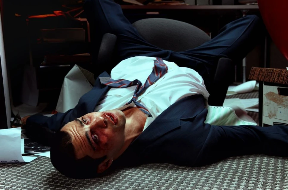 Injured adult male in a suit lies on a patterned floor, supported by an office chair, in an indoor environment with visible filing boxes.