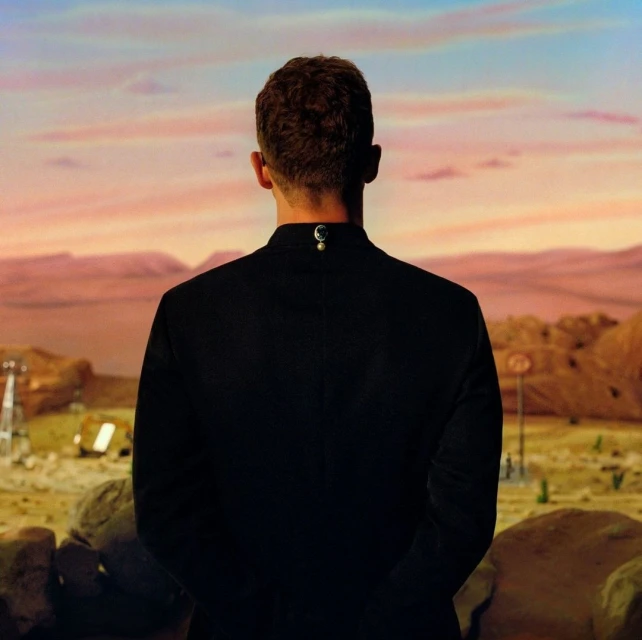 Man in black suit stands against a surreal desert landscape with orange sky and rock formations, creating a cinematic view.