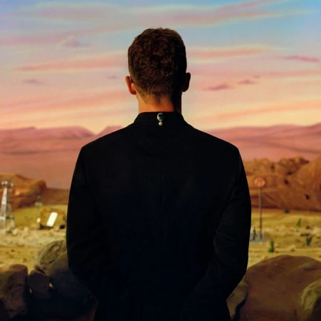Man in black suit stands against a surreal desert landscape with orange sky and rock formations, creating a cinematic view.