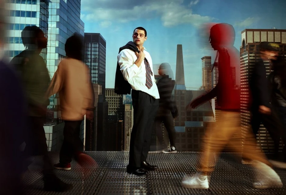 Man in dress shirt and trousers stands on a rooftop in a city with blurred pedestrians, holding his jacket over his shoulder.