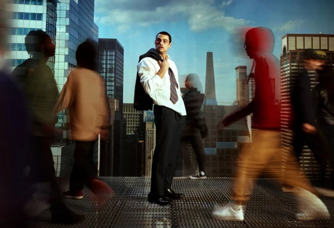 Man in dress shirt and trousers stands on a rooftop in a city with blurred pedestrians, holding his jacket over his shoulder.