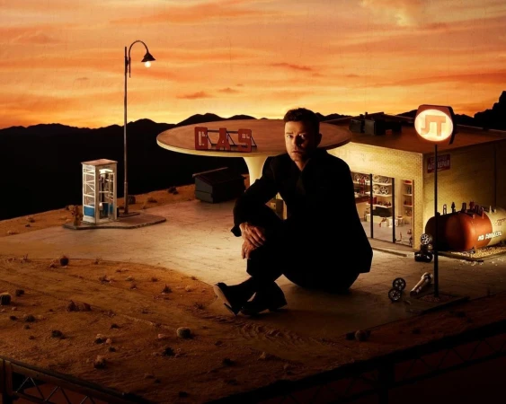 Adult male in black suit sits in front of miniature desert landscape with gas station at sunset. Miniature car and phone booth.