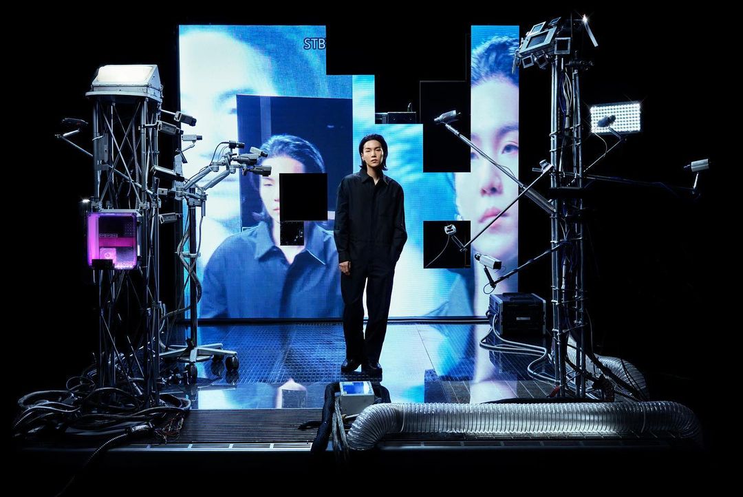 Man in black outfit stands on a platform surrounded by camera equipment, with a large screen displaying a blue-toned face behind him.