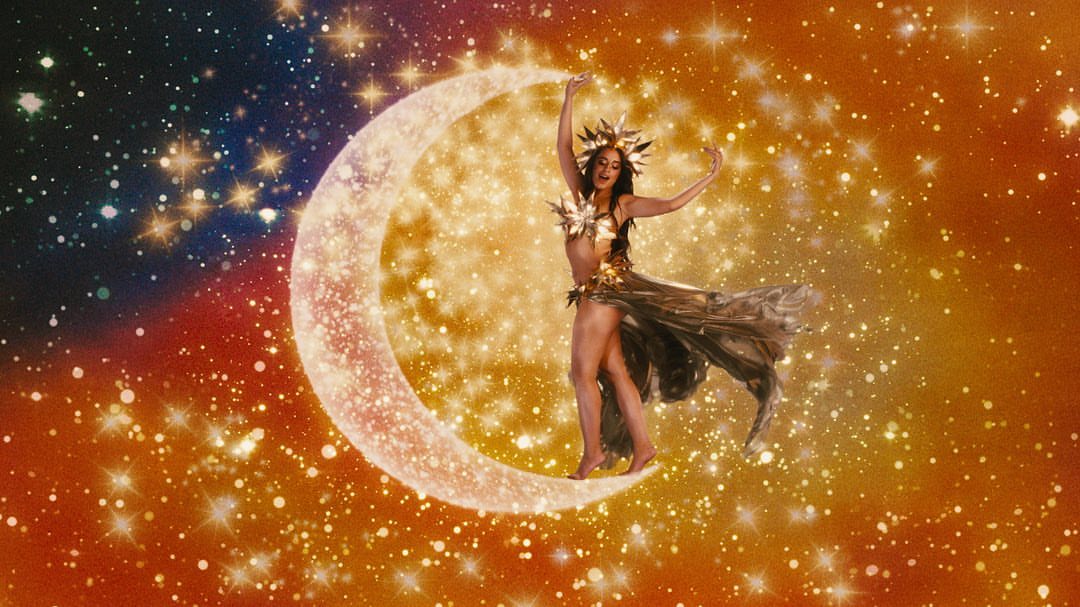 Woman posing on a crescent moon against a starry background, wearing a sun-shaped crown and ornate costume for a celestial performance.