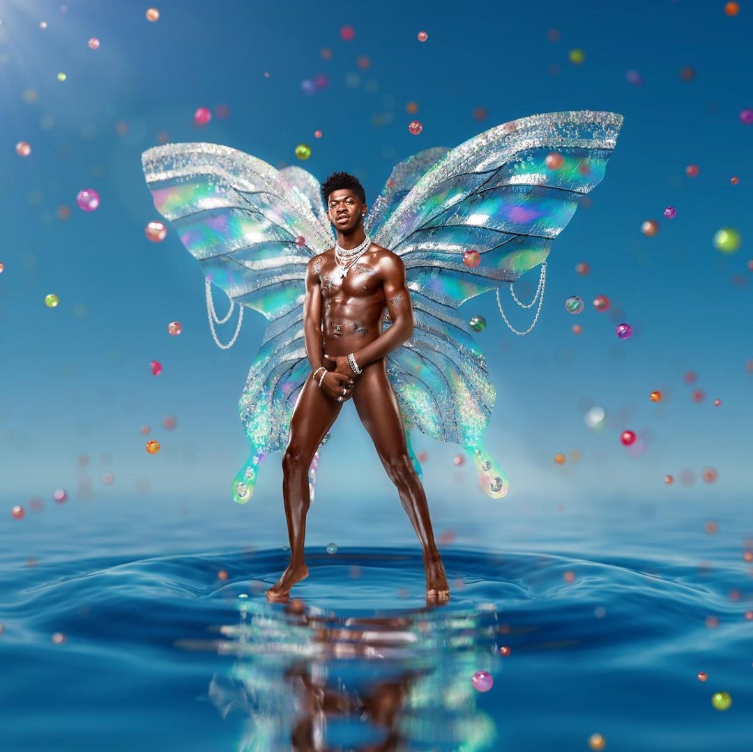 Lil Nas X poses in water with shimmering butterfly wings, wearing jewelry against a blue sky with floating spheres in the distance.