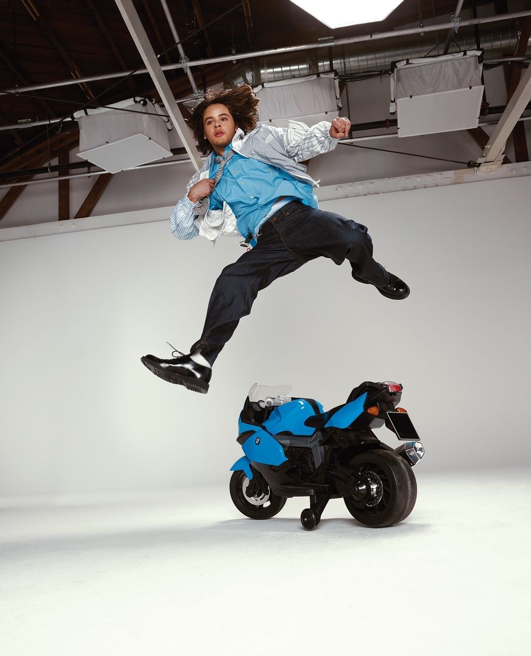 Young man mid-air near toy motorcycle in a studio. He wears a jacket, shirt, and jeans. Bright studio lighting illuminates the scene.