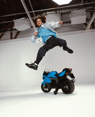 Young man mid-air near toy motorcycle in a studio. He wears a jacket, shirt, and jeans. Bright studio lighting illuminates the scene.