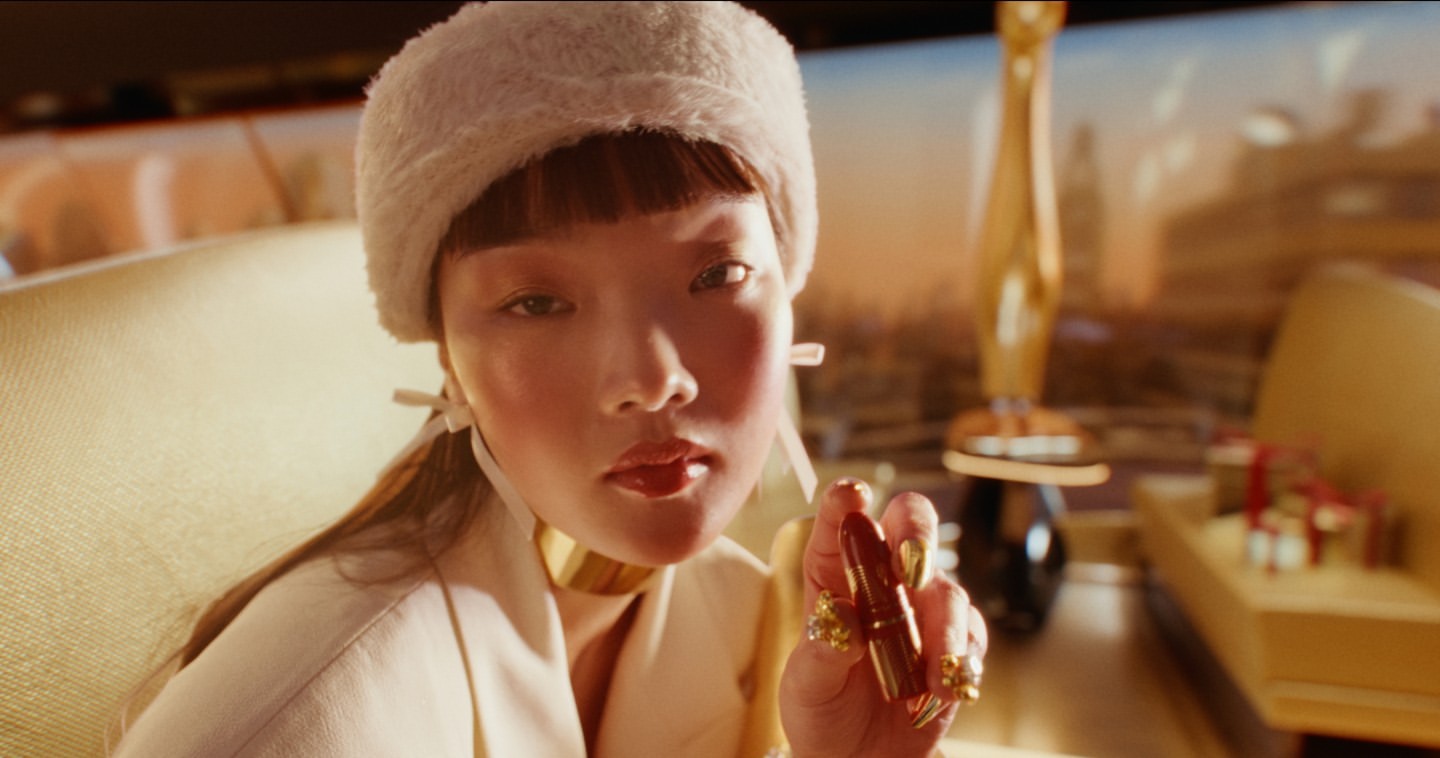Woman with gold jewelry, wearing a fuzzy hat, holding lipstick, indoors with blurred golden statue and gifts in background.