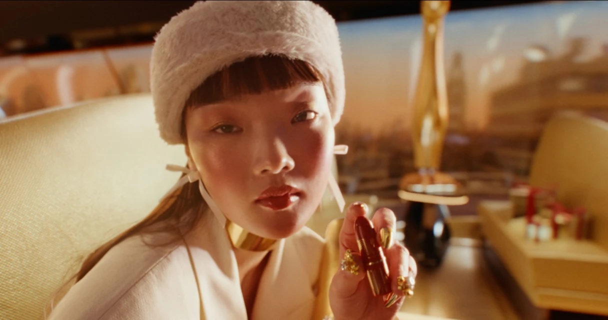 Woman with gold jewelry, wearing a fuzzy hat, holding lipstick, indoors with blurred golden statue and gifts in background.