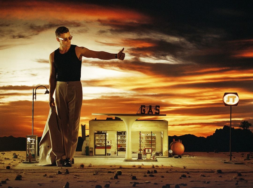 Adult male hitchhiking near a vintage gas station against a dramatic sunset sky in a stylized outdoor desert environment.