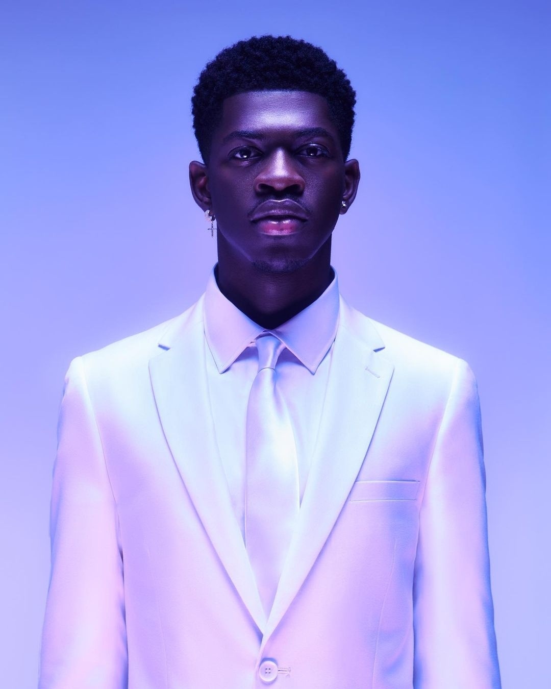 Close-up portrait of a young Black male in a white suit and tie, earrings, against a soft blue and purple gradient background.