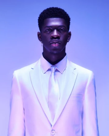 Close-up portrait of a young Black male in a white suit and tie, earrings, against a soft blue and purple gradient background.