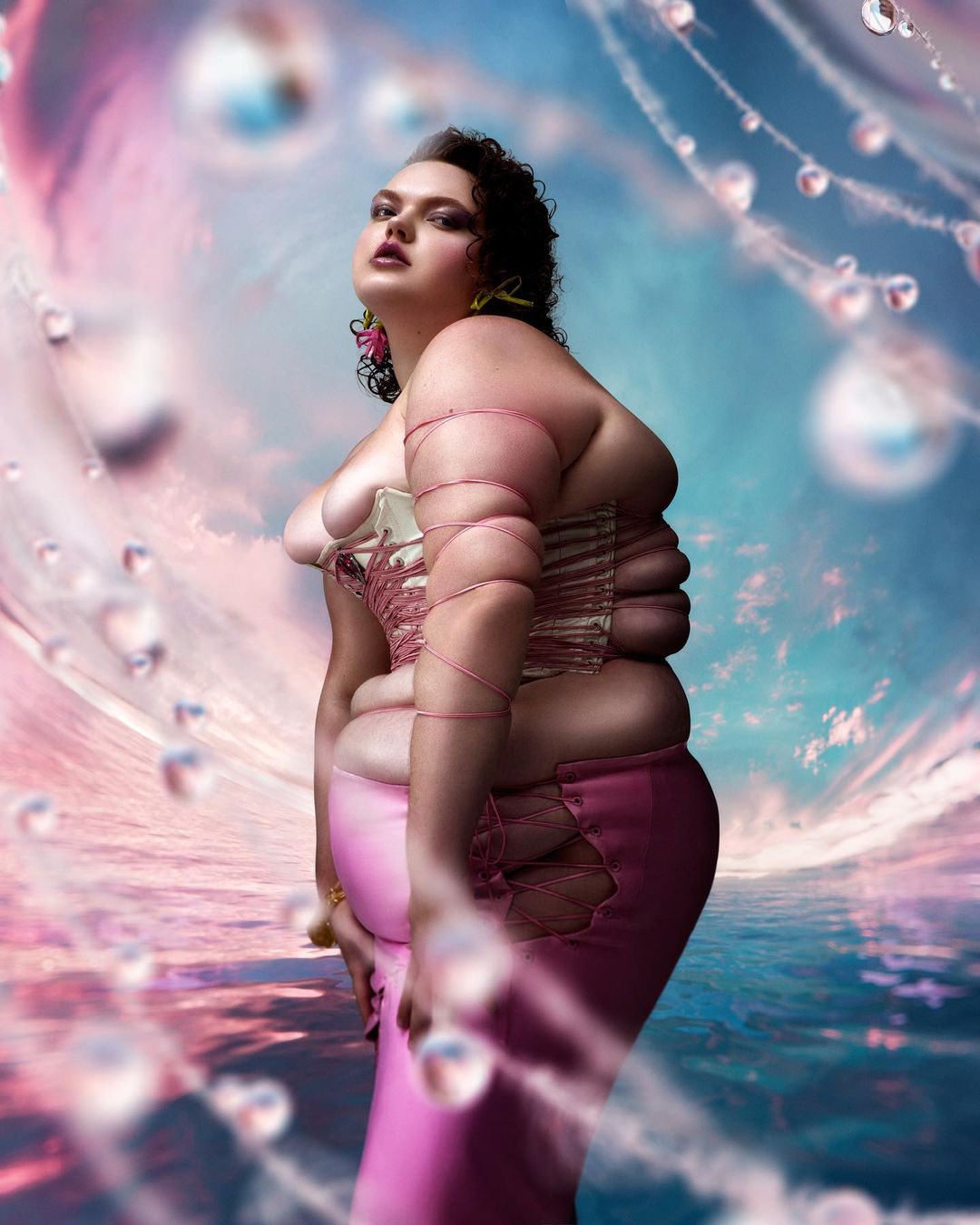 Woman poses in pink skirt and corset against an artistic backdrop with floating spheres, creating a surreal and eye-catching portrait.